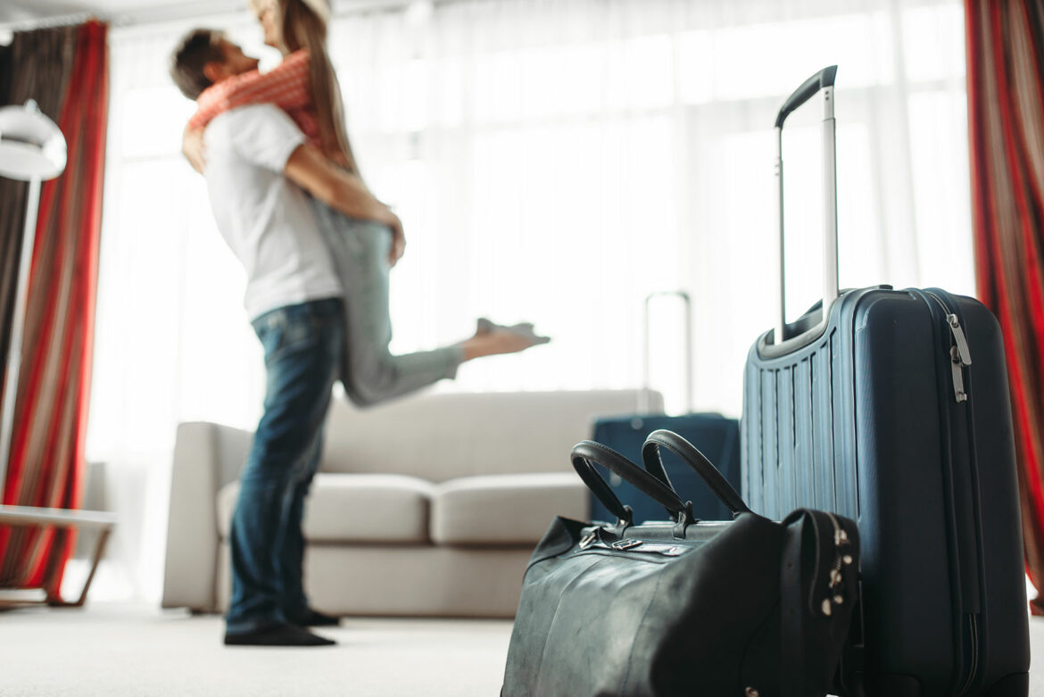 Hardside suitcase with handle extended and tote in front of an embracing couple