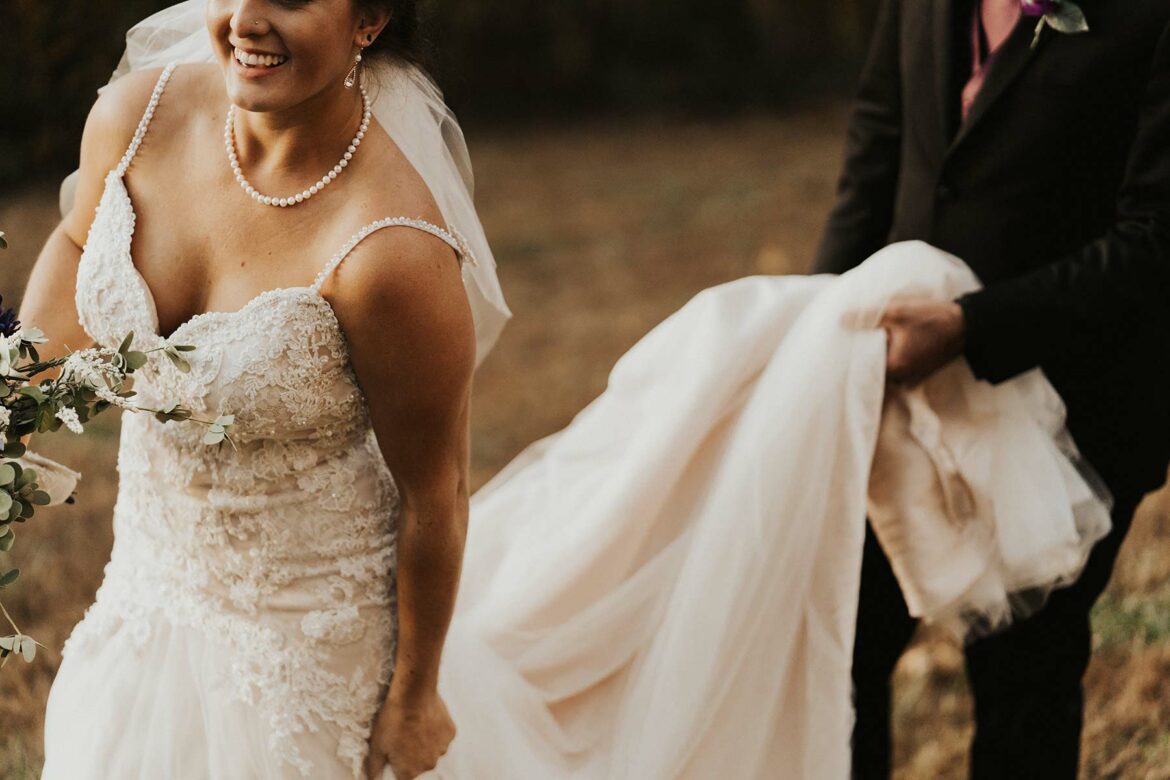 A close-up of a woman wearing a wedding dress and someone holding the train