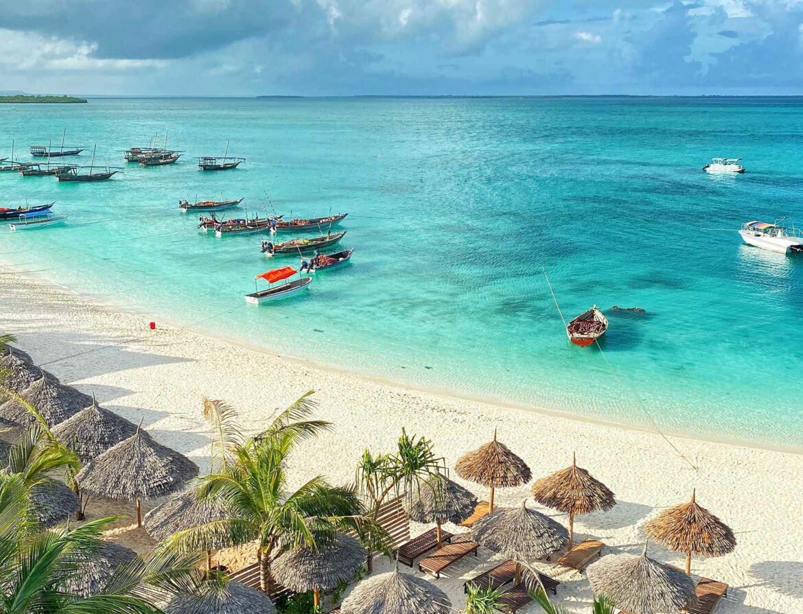 Palapas on a beach in Zanzibar and small boats in the blue water.