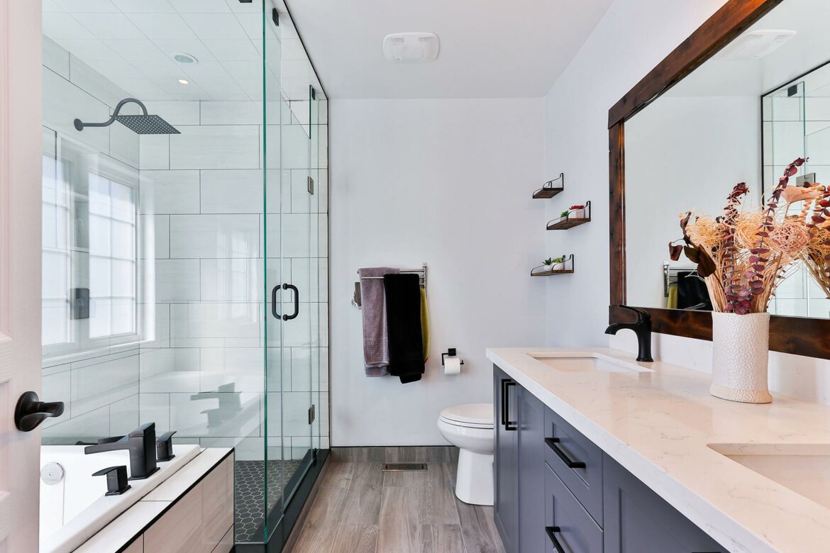 A bathroom with a glass shower and vanity with blue drawers and a white top. A vase filled with dried plants is on the vanity.