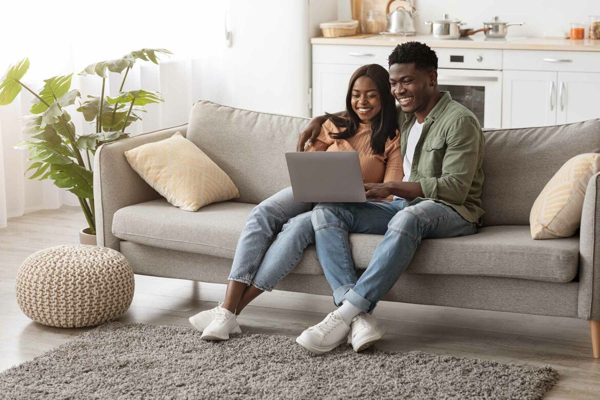 A couple sitting on a couch in a living room holding a laptop while creating a wedding registry