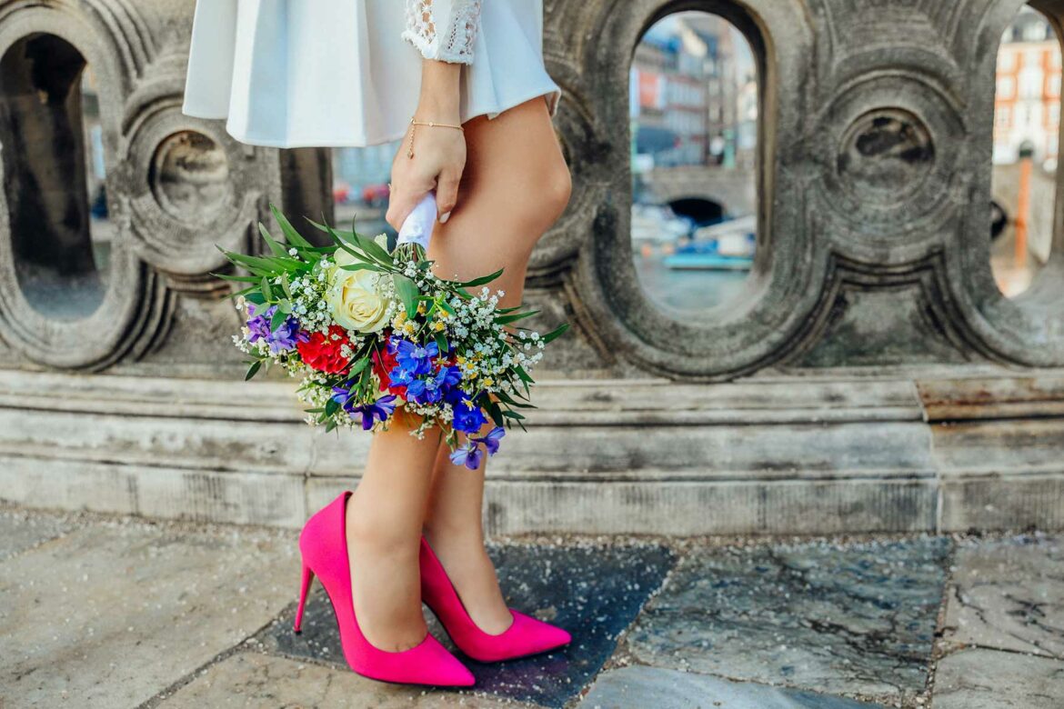 Closeup of a person holding a colorful flower bouquet and wearing white dress and hot pink pumps.