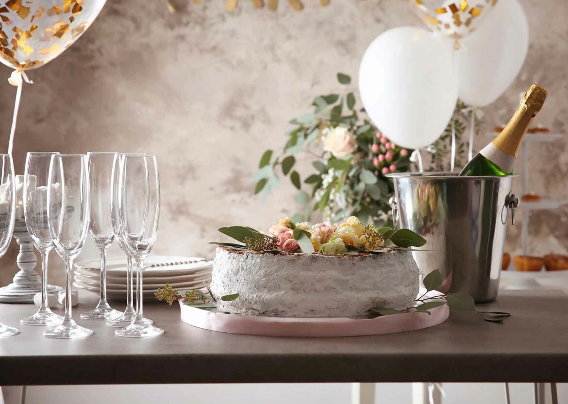 Wedding shower decorations including gold foil and white balloons, champagne in an ice bucket, glassware and a rustic cake on a wooden table